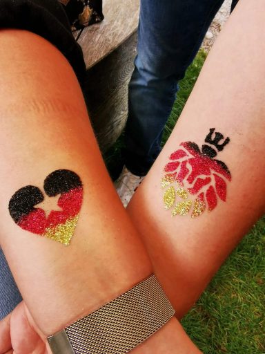 Zwei Tattoos auf Unterarmen: ein Herz in Schwarz-Rot-Gold und eine rote Hand mit Krone.